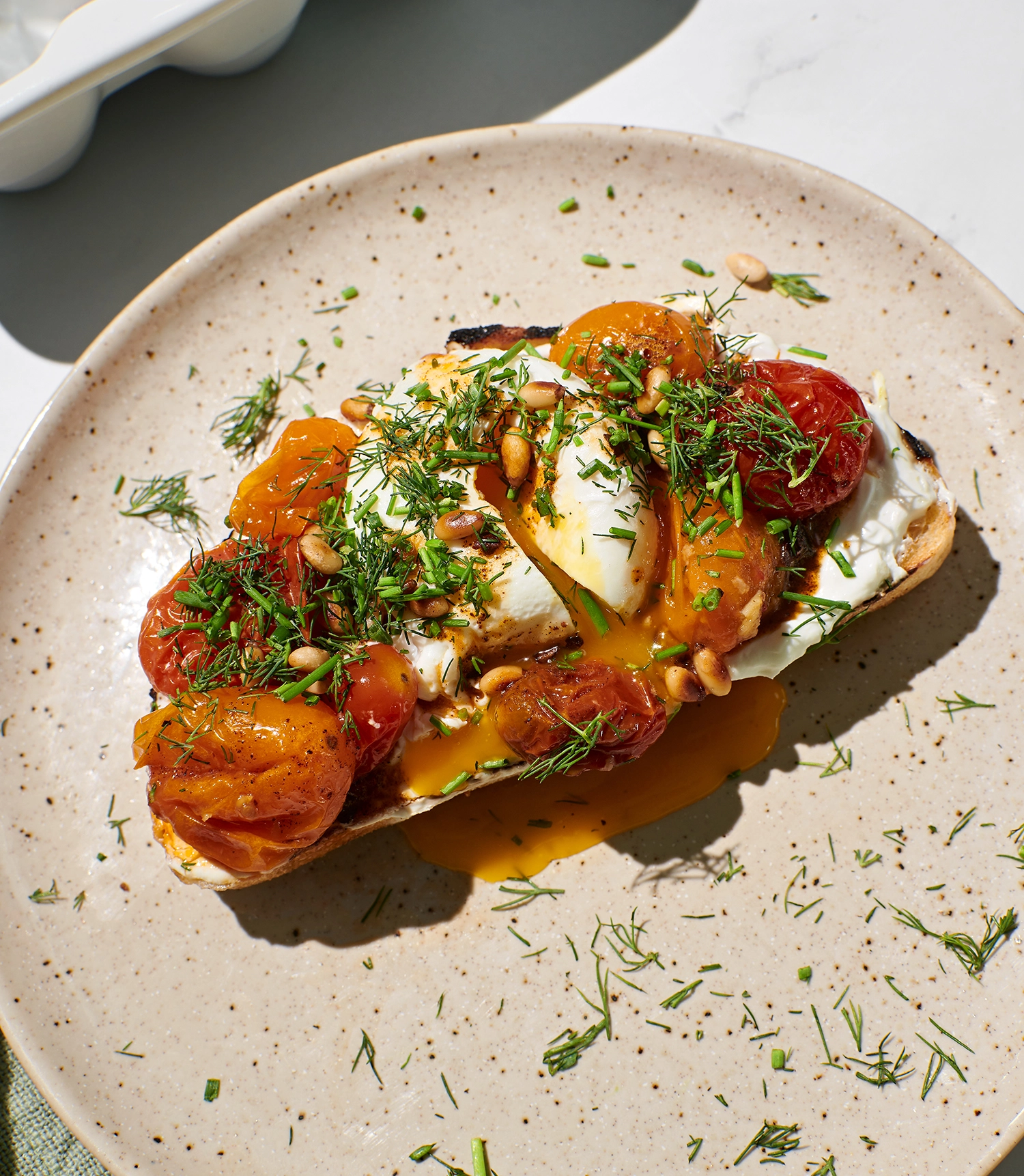 Garlicky Greek yogurt spread onto toast topped with roasted tomatoes, poached eggs, butter sauce, and garnished with toasted pine nuts, fresh chives, and dill served on a plate.