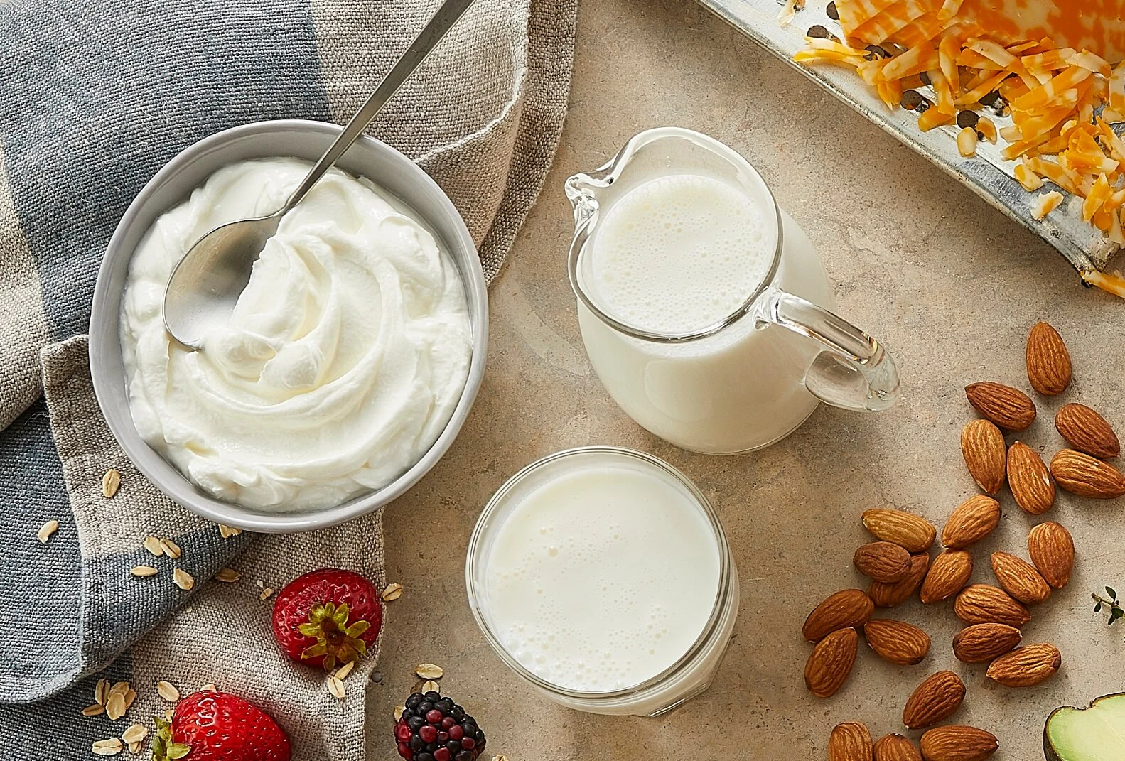A bowl of Greek yogurt with a spoon on a napkin next to a jug, a glass of milk, grated cheese, almonds, and strawberries.