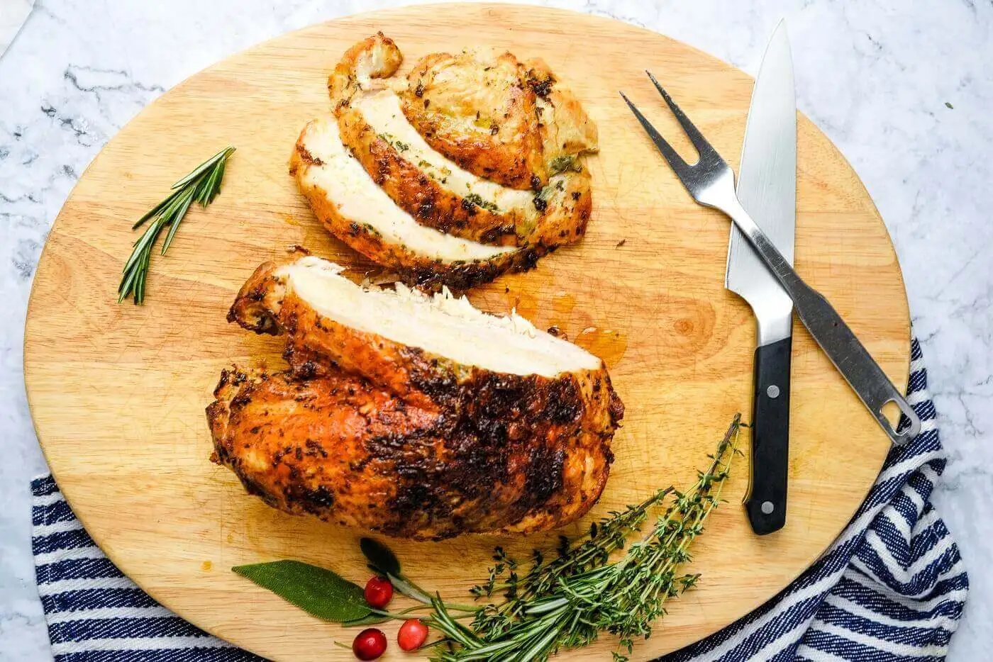 Slices of roasted turkey breast on a wooden cutting board with rosemary, thyme, and cranberries.