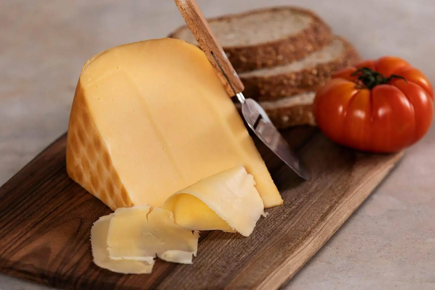 Wedge and thin slices of Gouda cheese on wood board next to wheat bread slices and a red tomato
